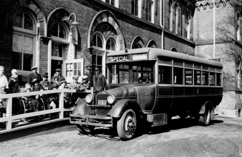 Children are lined up beside a bus. Some of them are sitting in wheelchairs. Two TTC drivers are standing beside the bus.