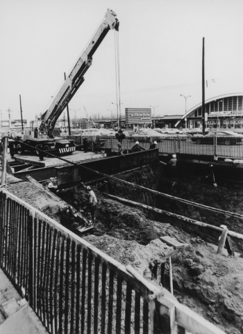 Crane and workers at excavated pit. Cars and mall in background.