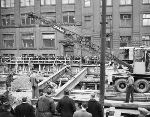 A crane lowers a large metal beam over an excavation, while workers guide the beam into place.