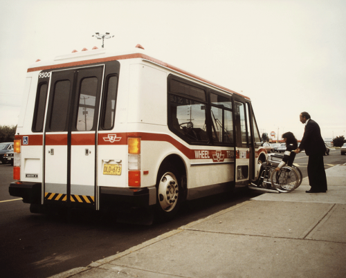 A woman sitting in a wheelchair rolls it up a ramp in front of a bus door. A man stands behind her with his hands on the handles of the chair.