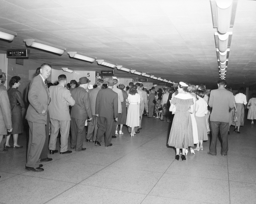 People are standing in lines in a long room with rows of lights on the ceiling.