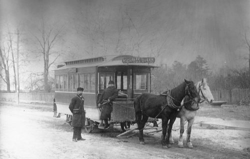 A streetcar pulled by a black horse and a white horse. A man in a winter coat and hat is standing on the step of the streetcar. The sign on the front says "High Park via Queen."