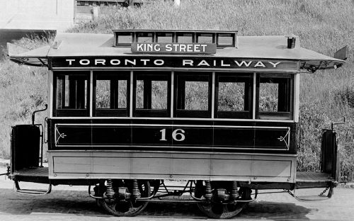 A small streetcar with six windows on each side, and four wheels. It is painted black and white. The sign on the roof says "King Street" and "Toronto Railway."