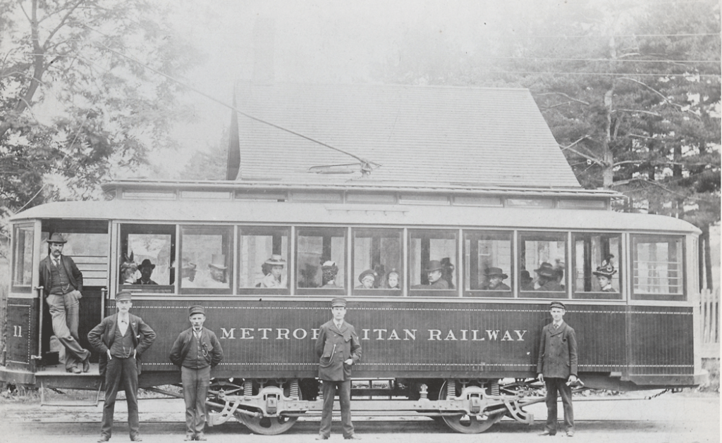 An electric streetcar with large windows. There are passengers inside looking out, and several men in drivers' uniforms are standing in front.