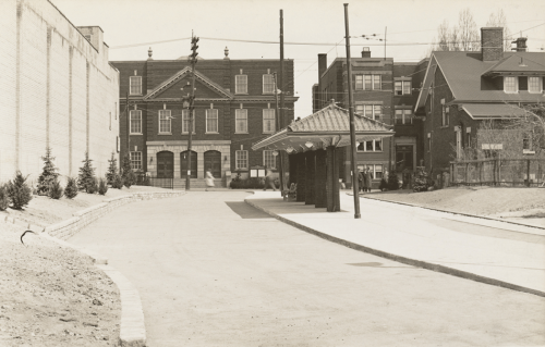 Vacant bus shelter with pillars, roof, and bench. Background shows houses and three storey brick buildings.