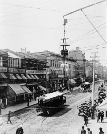 Streetcars drive down a street lined with brick buildings containing stores.