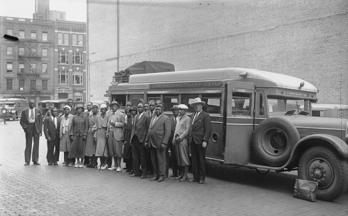 A group of Black people stands in front of a small bus. Behind them is a large brick wall probably the side of a theatre. In the background is a downtown street with stores.