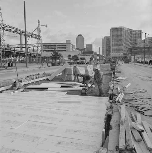 Four men laying down foam panels in the excavated middle lanes of the road. Background shows office towers and condominiums.