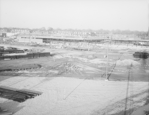 Foreground shows cleared area with rails for track installation. Background shows construction of large building.