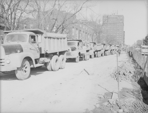 Large line of dump trucks on closed portion of road with traffic on the other side of the street.