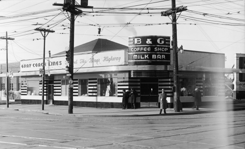 Exterior of a restaurant on a street corner. On the corner is a large curved sign reading "B and G Coffee Shoppe, Milk Bar." 