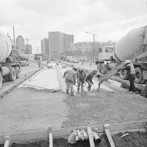 Four men laying concrete into prepared middle area of the road. Background shows office towers and condominiums.