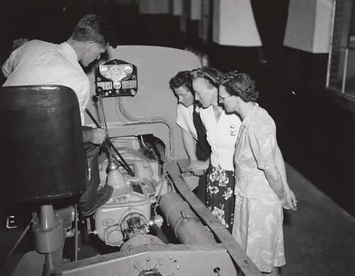 Three women looking at a bus motor with instructor pointing at motor part.