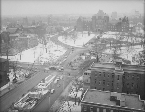 Winter scene showing streetcar, cars, and excavated area for tunnel construction. Foreground shows multi-storied building and background shows Parliament Buildings