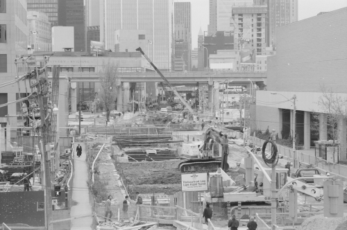 Installation of beams above excavated area. Foreground shows people on pedestrian passageway. Background shows office towers.