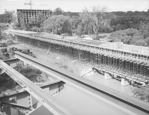 Installation of structural supports for bridge. Background shows trees and erection of structure for building.