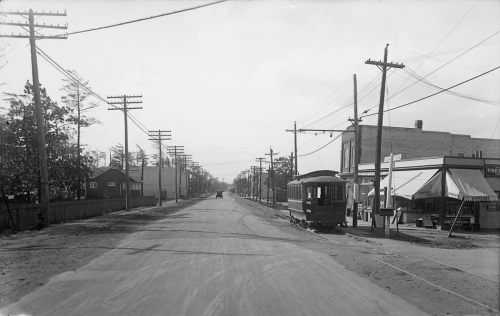 A streetcar drives along a wide road. On the right is a row of stores with large awnings.