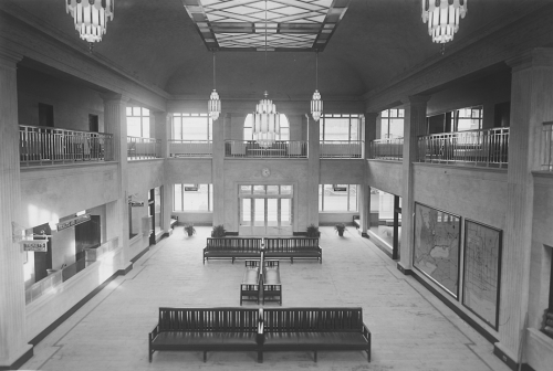 A waiting room that is two storeys high, with a stained glass skylight and large hanging chandeliers. The walls contain ticket windows and framed maps. There are several benches in the middle of the room. 