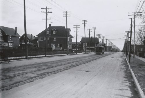 A streetcar drives down a street lined with telegraph and electricity poles. There are large houses in the distance.