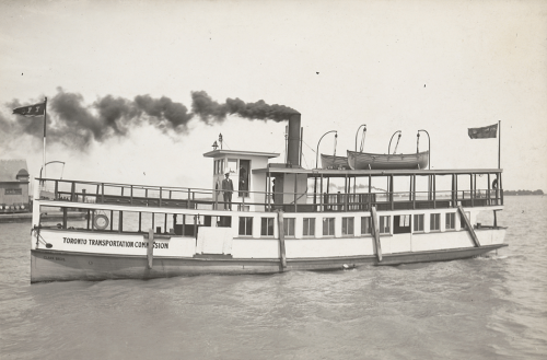 A wooden ferry with an enclosed lower deck and an open upper deck. There are two lifeboats on the top roof beside the smokestack.