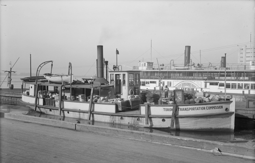 A wooden boat with its deck filled with boxes and parcels sits at a wide wooden dock.