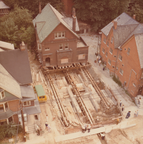 Three storey brick building removed from its foundation by rollers. House foundation can be seen with house roller mechanism.