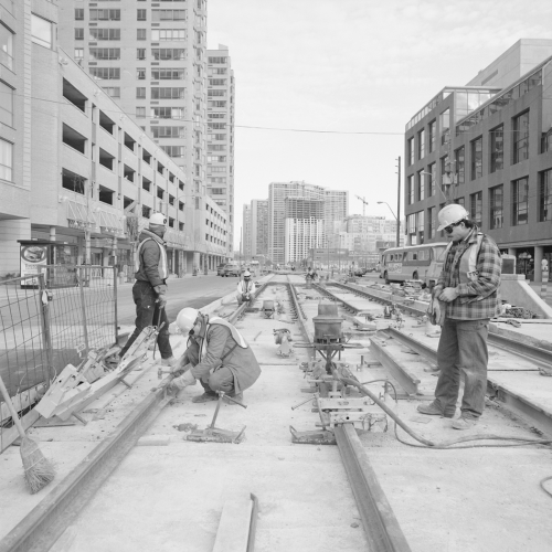 Three men installing streetcar rail in newly formed trackbed. Background contains office building, parking garages, and condominiums.