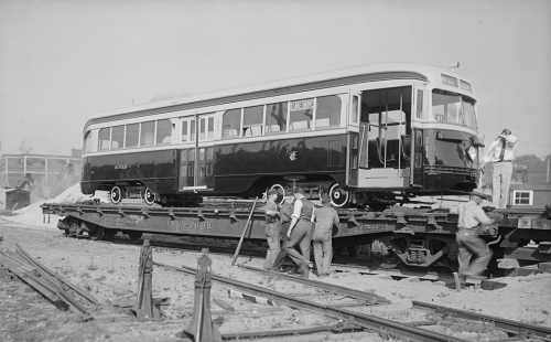 Workers are gathered around a shiny streetcar that sits on an open train car bed.