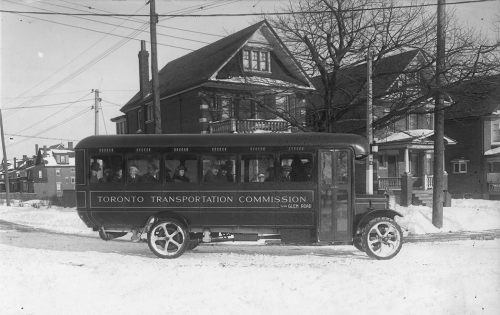 A small bus drives down a road in winter. There are detached three-storey houses behind it.