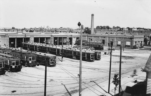 Several streetcars stand in a row outside of a low brick building with large garage doors.