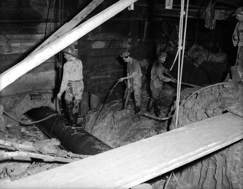 Workers are standing on a pipe and in a torrent of water in an underground excavation. 