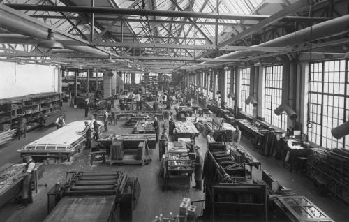 Interior of a large factory with big windows. Workers are assembling the stops of streetcars.