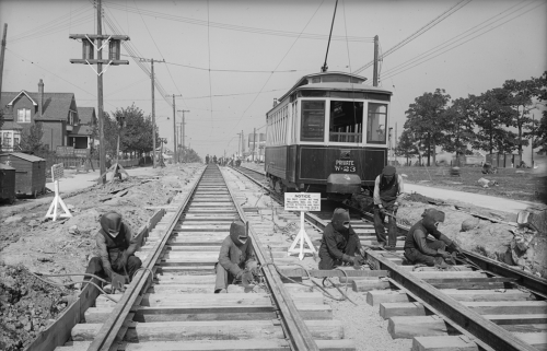 A row of men wearing metal welding helmets are putting together two sets of streetcar tracks on a road.