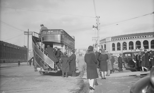 People in winter coats are boarding a double-decker bus with an open top. In the background is the outside of a two-storey sports stadium with large arched windows.