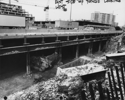 Cross section of tunnel construction under Yonge Street, cars moving above excavated area, and construction Of North York Civic Centre.