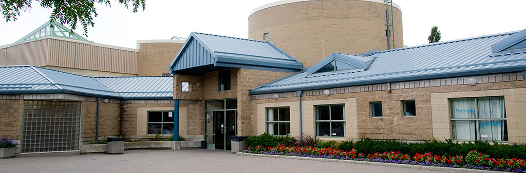 The exterior of Goulding Community Centre which shows the entrance, blue metal roof structure and planting beds with colourful flowers lining the exterior.