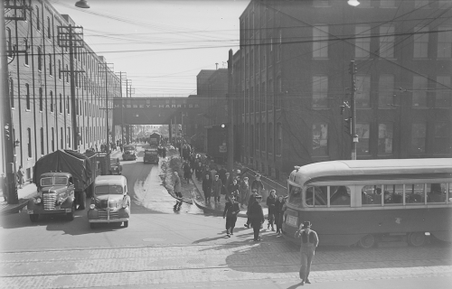 Crowd of people going to streetcar. Background shows large industrial brick buildings.