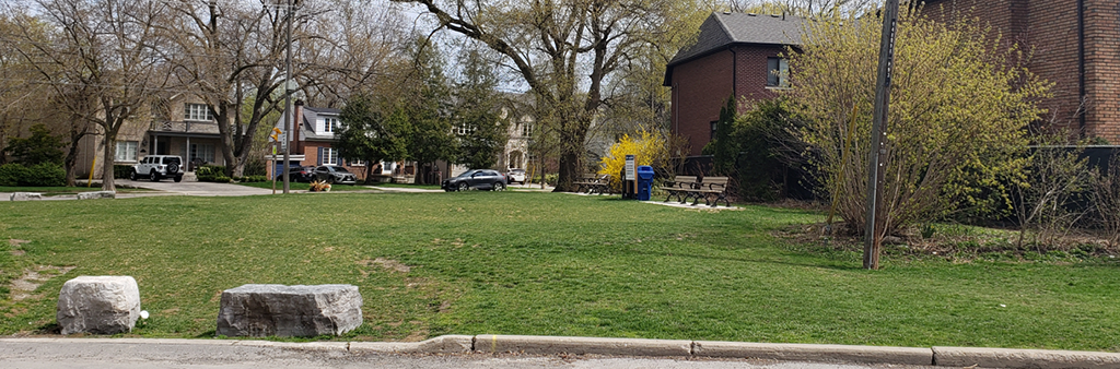 An image of the location for the new parkette which shows an open lawn space with two large boulders, four park benches, and residential homes with mature trees surrounding the site.
