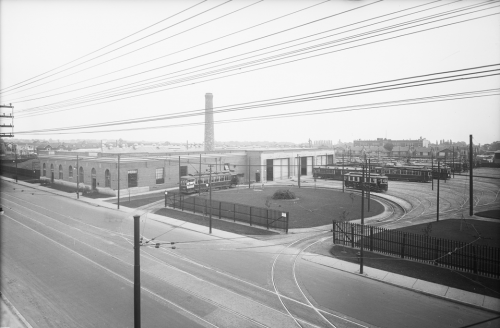 A low brick building with a large smokestack in the back. A streetcar is driving on a loop of track surrounding a grass lawn in the front.