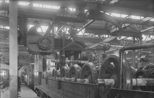 A worker in the bucket of a crane puts large metal wheels onto the bed of an open train car.