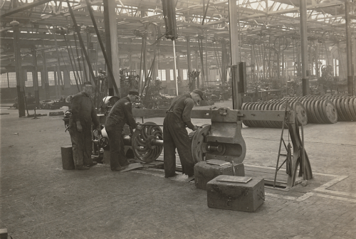 Workers are fitting metal wheels on to a small machine. Behind them is a large warehouse filled with metal wheels.