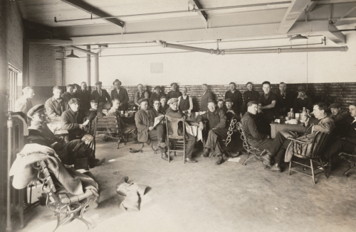 A group of men sit at tables and on wooden chairs and benches in the corner of an industrial-looking room. 