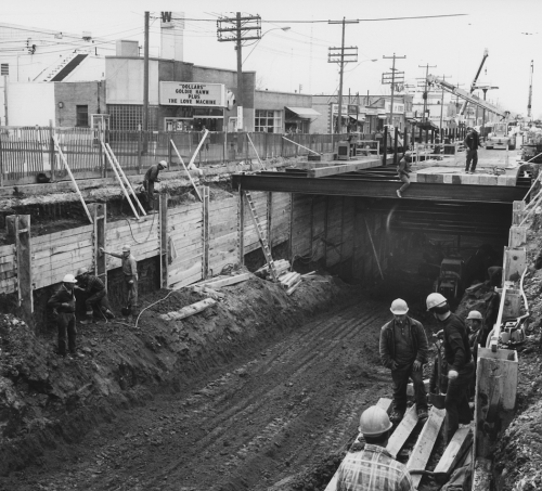 Cross section of mean and excavator in tunnel with beams on roadway. In the distance in heavy machinery.