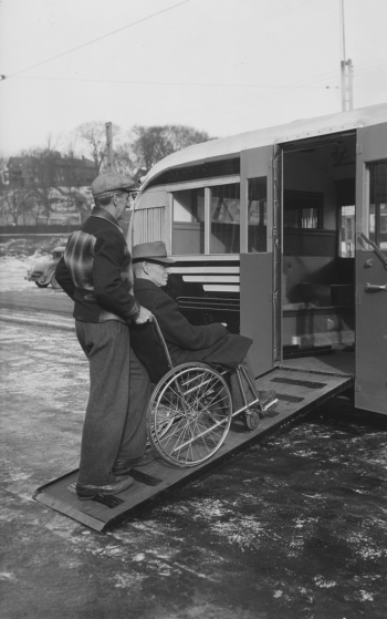 A man is sitting in a wheelchair. Another man is pushing the chair up a wooden ramp into a bus.