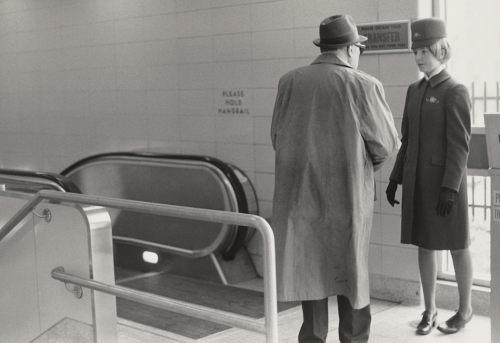 Uniformed woman with older man in trench coat standing at the top of an escalator.