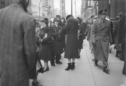 Crowded bus stop with woman offering assistance to woman buying TTC tickets.