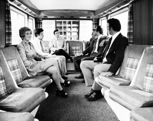 People are sitting inside a van that has wood panelling, plaid curtains, and bucket seats with paid accents facing across the aisle.