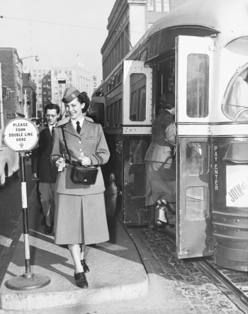 Uniformed women posing for the camera and offering a fare beside a streetcar.