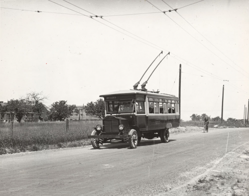 A bus is on a country road. There are metal arms coming up from the top of the bus to meet the electric wires overhead.