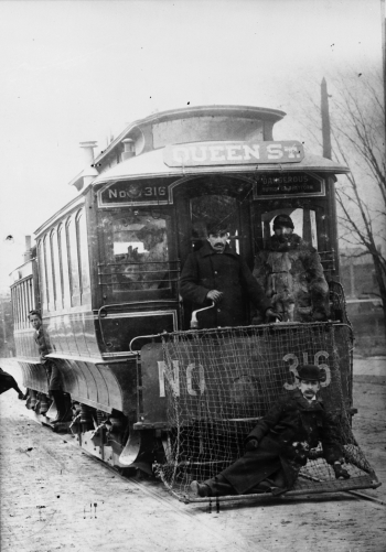 The front of a streetcar. A a man is lying on on the net attached to the front. The driver stands on an outdoor platform at the front of the streetcar.